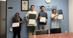 Four young people (two male and two female) hold up their Certificates of Completion in front of a wall with the Jay Nolan Community Services logo on it.