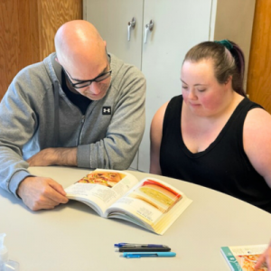 A man and a woman are seated at a table, reading a cookbook together, with two pens and a hand sanitizer bottle nearby.