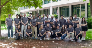 A group photo of the Twin Science team standing and sitting in two rows outdoors in front of a modern building. The team members, wearing matching gray T-shirts with the Twin Science logo, are smiling and posing together under the shade of a tree, with na