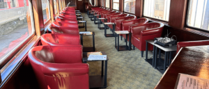 An empty scenic train car featuring rows of red leather chairs with small side tables, illuminated by sunlight streaming through large windows.