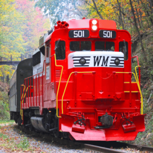 A bright red train with the label "WM" on the front, traveling through a forested area with autumn-colored trees in the background.