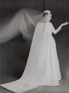 Black and white artistic photo of a woman with her right arm bent wearing a structured white wedding dress and veil floating in the air