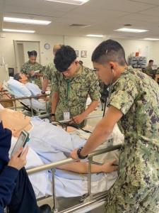 Sea Cadets participating in medical training in a hospital setting. Two cadets in camouflage uniforms are practicing medical procedures on a mannequin patient in a hospital bed. One cadet is using a stethoscope, while the other is adjusting equipment.