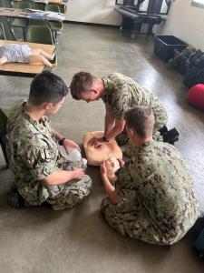 Three Sea Cadets in camouflage uniforms practicing CPR on a training mannequin. One cadet is performing chest compressions, while another is holding the mannequin’s head to maintain an open airway, and the third cadet is preparing a bag valve mask for ven