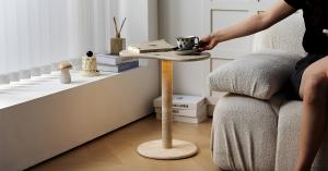 A modern PeaPod Travertine Side Table by OIXDESIGN, featuring a natural stone surface, with a cup of coffee and a book on top, showcasing its practicality and elegance in a cozy living room setting.