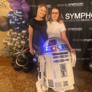 Two individuals smiling and posing with an R2-D2 replica in front of a Sequoia Symphony backdrop, surrounded by purple and silver balloon decorations.