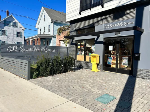 The storefront of All She Wrote Books, featuring the name of the store in cursive script, an outdoor area with patio and greenery, as well as a bike rack and LGBTQIA+ memorabilia in the bookstore windows.