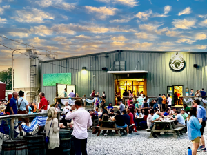 A photo of a one story building, WISEACRE's Broad Avenue taproom, with dozens of people standing outside of the taprooms patio enjoying beers.