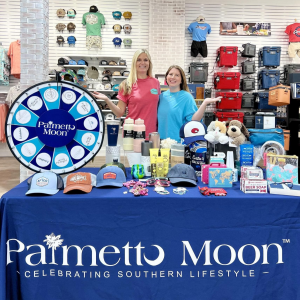 Two Palmetto Moon employees stand in front of a spin-to-win wheel with prizes that are available to win at the Grand Opening Event.