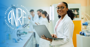 Researcher in a lab holds laptop, looks at the camera