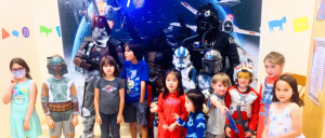 Children dressed in costumes pose in front of a space-themed backdrop with characters from a popular sci-fi universe.