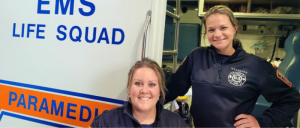 Two paramedics in uniform standing beside an ambulance with "EMS LIFE SQUAD PARAMEDIC" text.