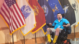 Sgt. Jordan Teegardin, a wheelchair user, poses at Shades of Green in front of multiple military flags, including the American flag and flags representing various branches of the U.S. Armed Forces. He is wearing a blue shirt, light-colored shorts, and yel