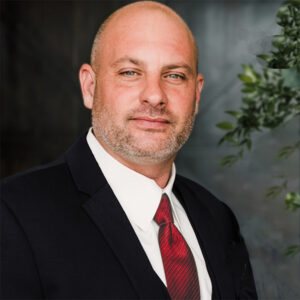 Portrait of Ken Wells wearing a suit and red tie, representing his leadership roles at Coryell Roofing.