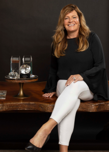 A woman in black and white sits on a table next to glass awards and a black background.