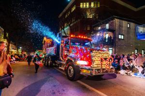 A truck covered in bright Christmas lights driving down State Street in Kennett Square during a holiday parade. A snow machine sprays artificial snow into the crowd that is gathered to watch the parade.