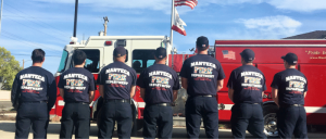 Firefighters from Manteca Fire Department standing in front of a fire truck, facing the American flag.