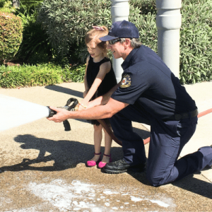 A firefighter is kneeling and helping a child hold a fire hose, spraying water on concrete.