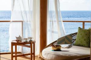 A tranquil setup featuring a tea set and light snacks on a wooden table, placed near a daybed with green cushions, overlooking a sunlit ocean framed by sheer white curtains.