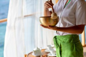 A close-up of a staff member holding a golden singing bowl with a mallet, set against a backdrop of soft white curtains and a tea setup overlooking the ocean.