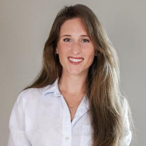 A professional headshot of a smiling woman with long, light brown hair. She is wearing a white collared shirt and a delicate cross necklace, standing against a neutral gray background.