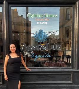 Pamela Maria De La Luz at Last Chance for Animals’ 40th Gala, standing in front of the bar with the AgaveLuz Organic Tequila logo displayed on the entrance window.