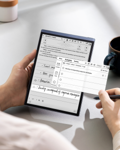 A hand holds the AINOTE Air 2 smart notebook, which rests on a table. The screen displays a mix of handwritten notes and typed text, along with icons for various functions. A stylus is used to write on the screen, demonstrating the device's note-taking ca