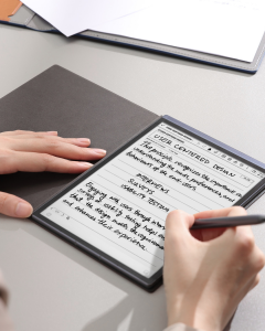 A person uses a stylus to write on the AINOTE Air 2 smart notebook. The notebook displays handwritten notes and digital text