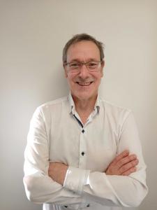 Portrait photo of Mark Hughes CCO, standing smiling in a white shirt.