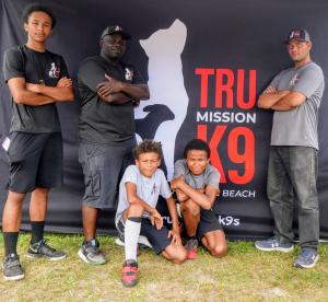 A family-style group photo of the Tru Mission K9s core team, featuring Don White, the founder, standing confidently with his arms crossed in the center. Beside him stands his eldest son, Josiah White. Kneeling in front are Jay and Joel White, alongside te