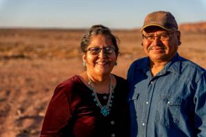 A smiling senior couple standing outdoors in a sunny, open desert landscape. The man is wearing a denim shirt and a cap, while the woman is dressed in a burgundy top with turquoise jewelry, representing a sense of togetherness and well-being.