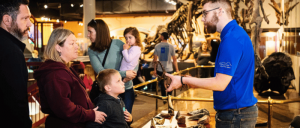 A museum staff member interacts with visitors near a dinosaur skeleton exhibit.