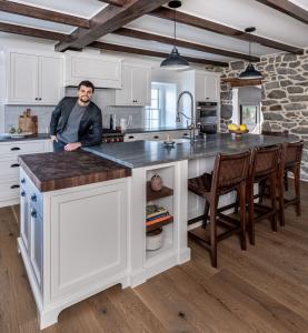 A man in black and gray clothing leans on a kitchen island with white cabinets and a gray countertop. A stone wall is to the right.