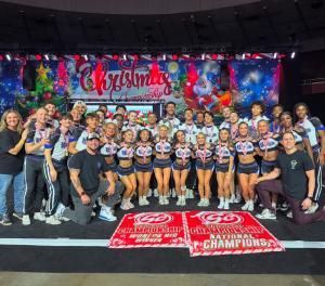 A large group of cheerleaders from Cheer Athletics’ Wildcats team pose together on a stage at the Christmas Championship event. The team is wearing matching blue, white, and sparkly uniforms, with medals around their necks. . Two red banners are displayed