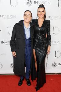 Photography of two women standing in a red carpet. On the left, Maria Gadu, a white, short and bald woman wearing blue suit and glasses. On the right, Thais Eliasen, white and tall woman, wearing a black and shiny dress. Her hair is up on a tall ponytail.