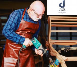 "An experienced craftsman wearing a leather apron works on a wooden chair frame with a power tool in the Dogan London workshop, showcasing precision and dedication to bespoke furniture making."