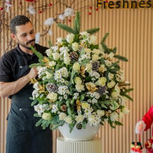 A florist arranges a stunning Christmas centerpiece with white roses, daisies, pine cones, and evergreens, set against a festive holiday backdrop.