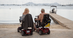 An elderly couple smiling by the water, enjoying the outdoors with their stylish 4-wheel mobility scooters, representing freedom and adventure in 2025.