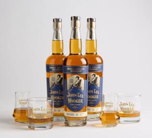 Three bottles of John Lee Hooker Legacy Spirits Boogie Chillen Bourbon. The label features an illustration of John Lee Hooker. Four glasses etched with John Lee Hooker logo glasses