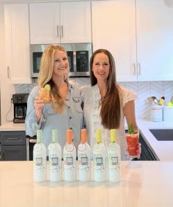 Sisters Jocelyn Dunn and Jordan Catapano stand in a brightly lit white kitchen with their white cocktail mixer bottles. On the left is Jocelyn, holding a Margarita and on the right is Jordan, holding a Bloody Mary.