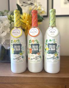 Three white bottles of non-alcoholic cocktail mixers are lined up in a row on a dark wooden table. A bouquet of flowers are just behind on the left and in the background are the bottom corners of two paintings.