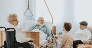 Elderly man in a medical bed at home holding a trapeze bar, surrounded by family members visiting and engaging in conversation.