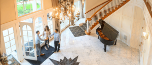 Elegant foyer with marble floor, grand staircase, chandelier, and three people greeting.