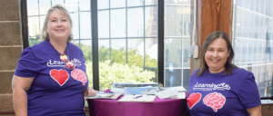 Two people in purple shirts standing behind a table with pamphlets, indoors near a window.