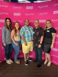 A group of five smiling individuals standing in front of a bright pink #GirlsClub banner with the text “Promote Badassery: Be Real. Be Authentic. Be You.”