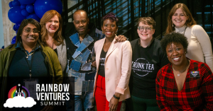 A group of seven individuals smiling in front of a festive backdrop with blue balloons, representing the Rainbow Ventures Summit.