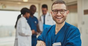 Man smiling and wearing blue scrubs in the foreground with out-of-focus team of medical providers collaborating in the background.