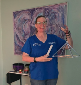 Dr. Couldry, Woman with blue scrub top and quartz pyramid with mallet, singing bowls in background and candles