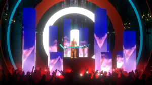 A vibrant virtual concert stage setup featuring a DJ booth with a glowing holographic performer at center stage, surrounded by tall illuminated blue rectangular pillars and a large white circular arch backdrop. The foreground shows silhouettes of a cheeri