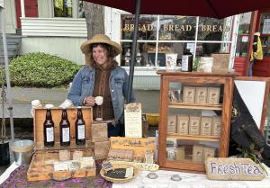 Shaelee Evans smiling at a farmers market booth under a red umbrella, surrounded by tea products and a rustic display.